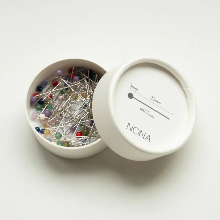A white round pin box shown open with colorful sewing pins inside, alongside its lid printed with size information. The brand name NONA is visible on the lid.