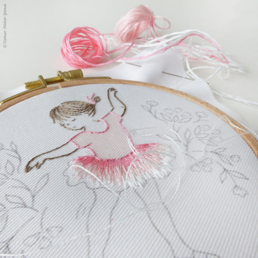 Close-up of the Little Dancer Embroidery Kit in progress, featuring detailed hand embroidery on white fabric with pink and brown embroidery floss and floral sketch outlines.