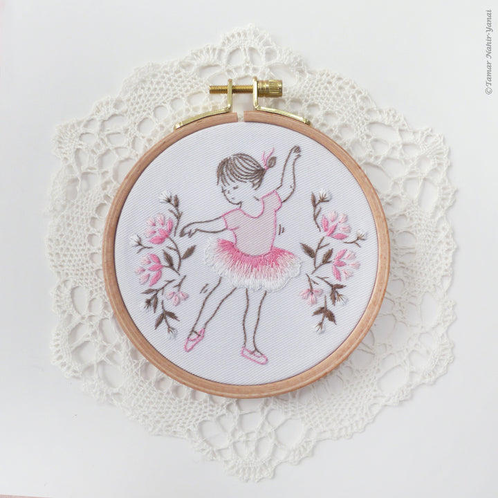 Little Dancer Embroidery Kit featuring a delicate ballerina in pink and white thread with floral accents, displayed in a wooden hoop on a lace doily backdrop.