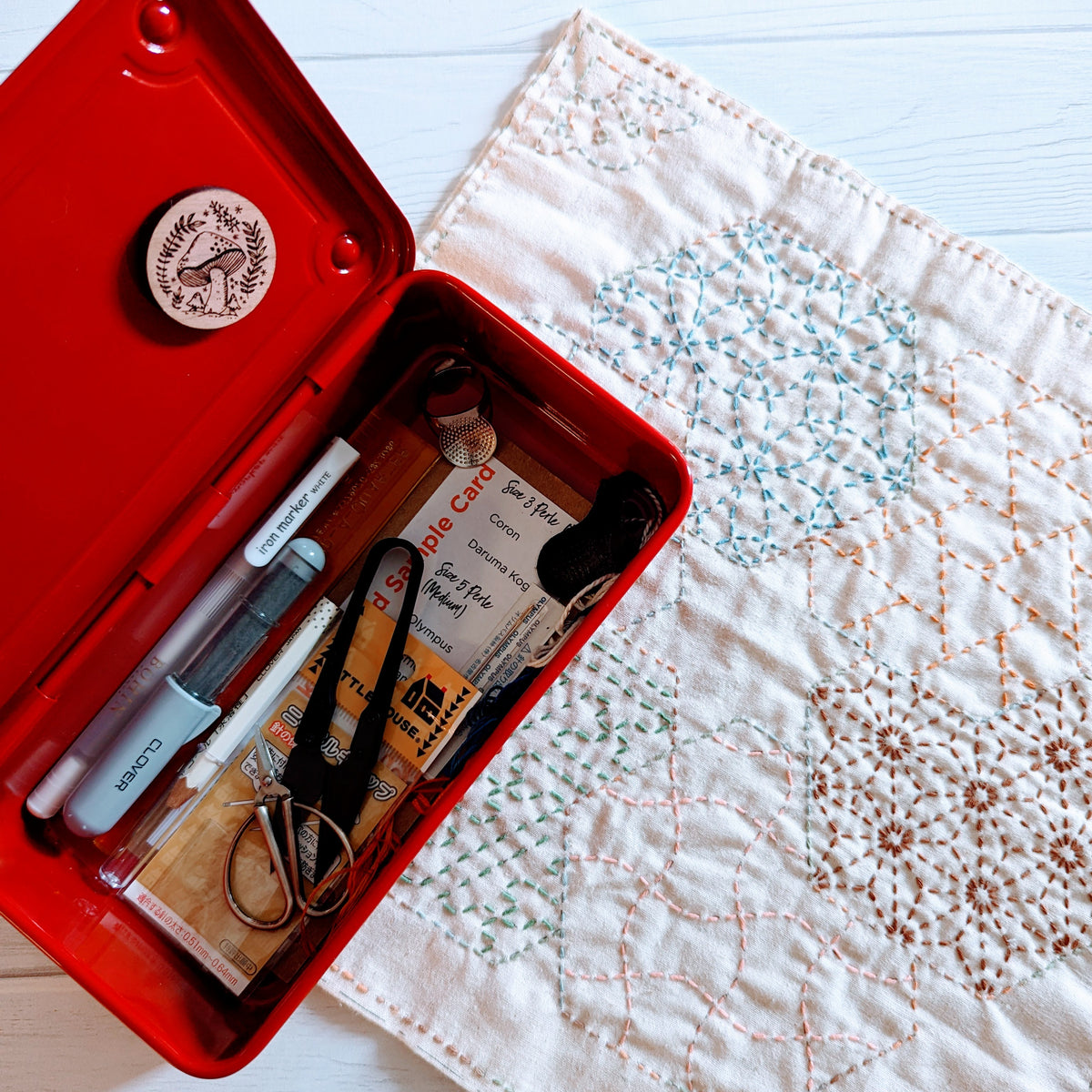 Toyo Steel T-190 Metal Storage Box open with embroidery tools inside, next to a white fabric square featuring colorful sashiko stitch patterns in blue, green, and brown.