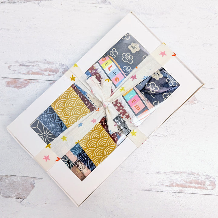 Boro Fabric Scrap Box (8 oz) featuring assorted Japanese fabric scraps in blue, yellow, and patterned designs, tied with a cream ribbon decorated with colorful stars.