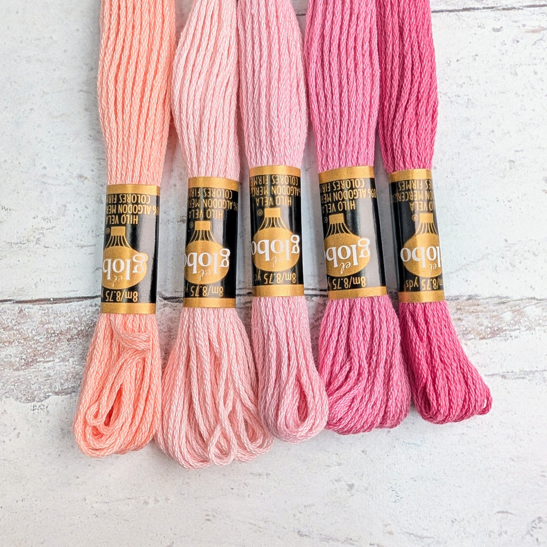 Five skeins of Hilo Vela El Globo Embroidery Thread (8m Skeins) - Light Pink shades arranged on a wooden surface, showcasing soft pink to deep rose tones for needlework projects.