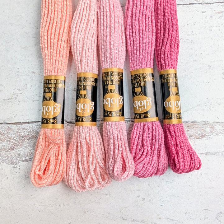 Five skeins of Hilo Vela El Globo Embroidery Thread (8m Skeins) - Light Pink shades arranged on a wooden surface, showcasing soft pink to deep rose tones for needlework projects.
