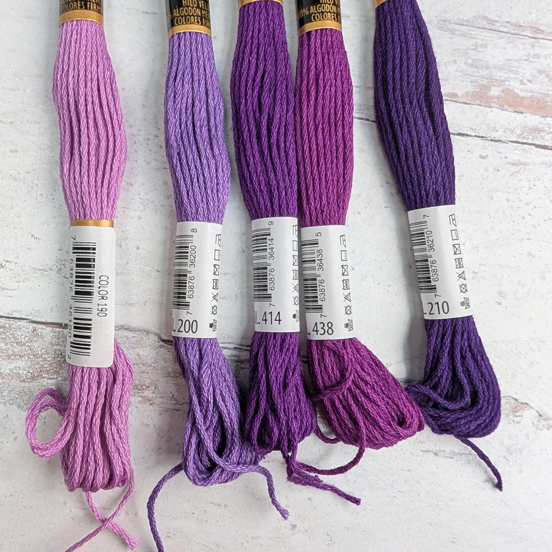Five skeins of Hilo Vela El Globo Embroidery Thread (8m Skeins) - Purple in shades ranging from light lavender to deep violet on a light wooden surface.