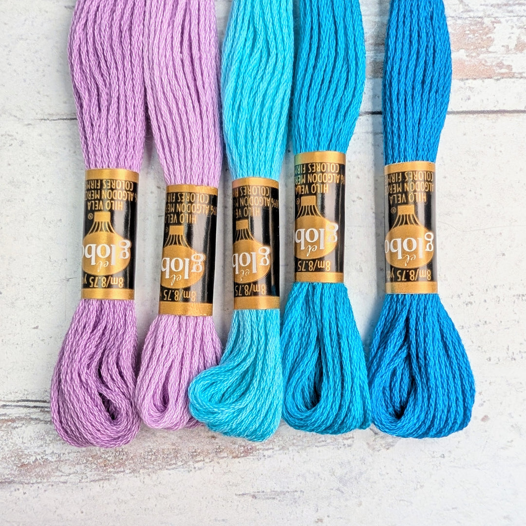 Five skeins of Hilo Vela El Globo Embroidery Thread (8m Skeins) - Lilac Pacific Blue in shades of lilac and blue arranged on a light textured surface, ideal for embroidery and needlework.