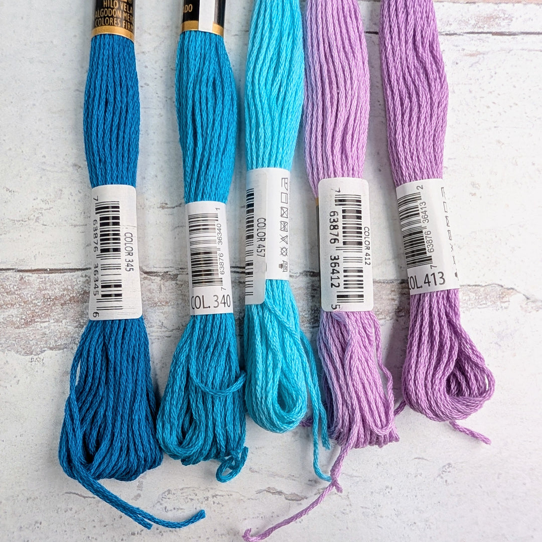 Five skeins of Hilo Vela El Globo Embroidery Thread (8m Skeins) - Lilac Pacific Blue in shades of bright blue, turquoise, and lilac arranged on a white textured background.