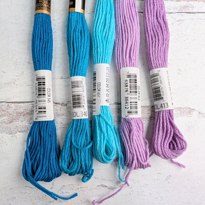 Five skeins of Hilo Vela El Globo Embroidery Thread (8m Skeins) - Lilac Pacific Blue in shades of bright blue, turquoise, and lilac arranged on a white textured background.