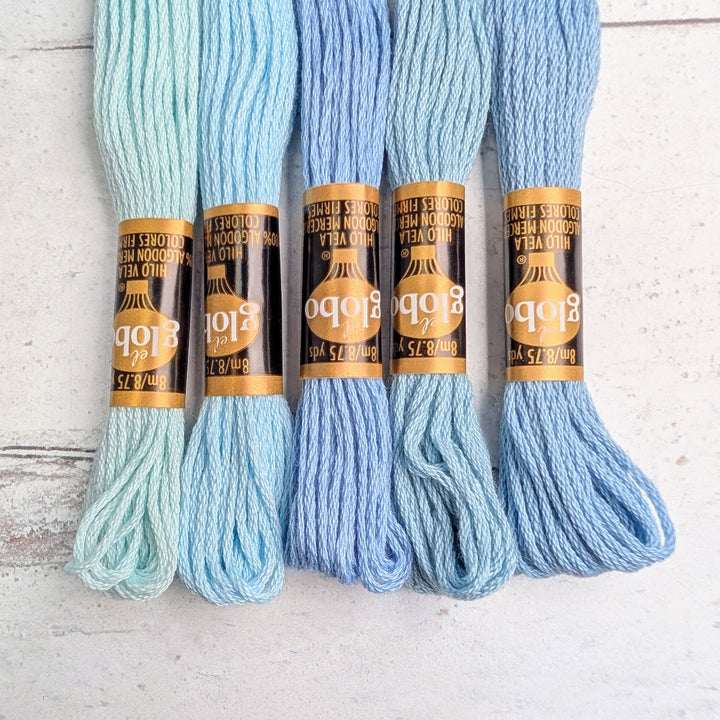 Five skeins of Hilo Vela El Globo Embroidery Thread (8m Skeins) in various shades of light blue arranged side by side on a white surface, showcasing high-quality needlepoint thread for modern embroidery projects.