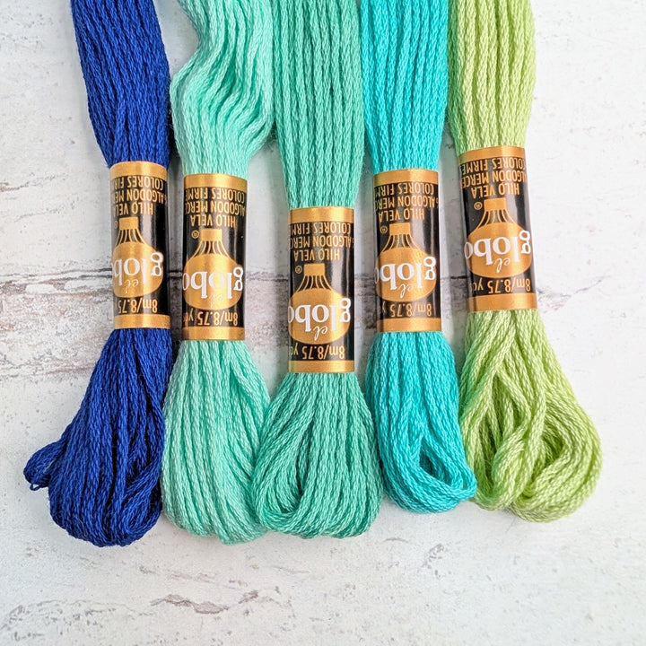 Five skeins of Hilo Vela El Globo Embroidery Thread (8m Skeins) -Blue Green in varying shades from dark blue to light lime green, arranged on a light textured surface.