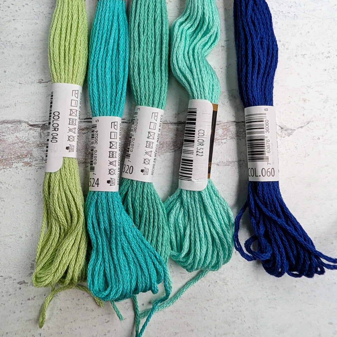 Hilo Vela El Globo Embroidery Thread (8m Skeins) -Blue Green in various shades of blue and green displayed on a rustic white wooden surface, showcasing vibrant embroidery floss for needlework and cross stitch projects.