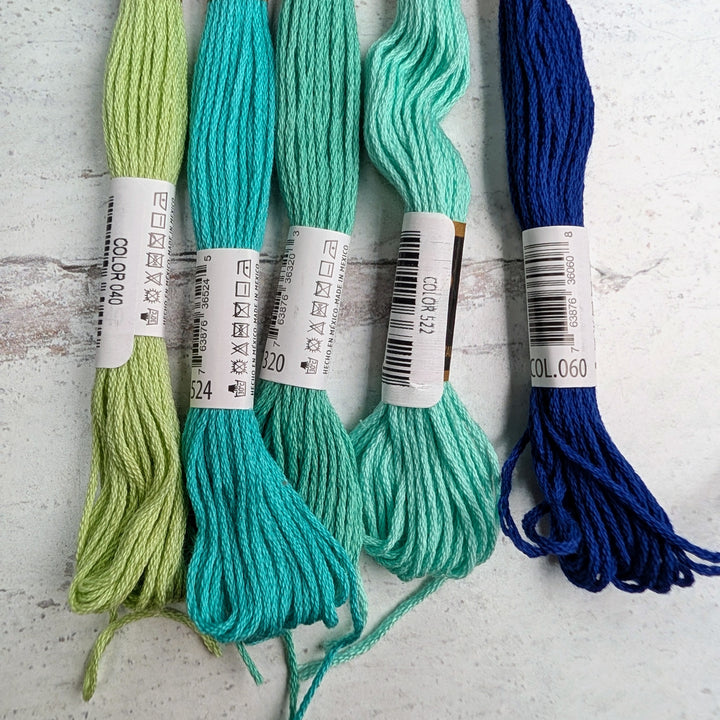 Hilo Vela El Globo Embroidery Thread (8m Skeins) -Blue Green in various shades of blue and green displayed on a rustic white wooden surface, showcasing vibrant embroidery floss for needlework and cross stitch projects.