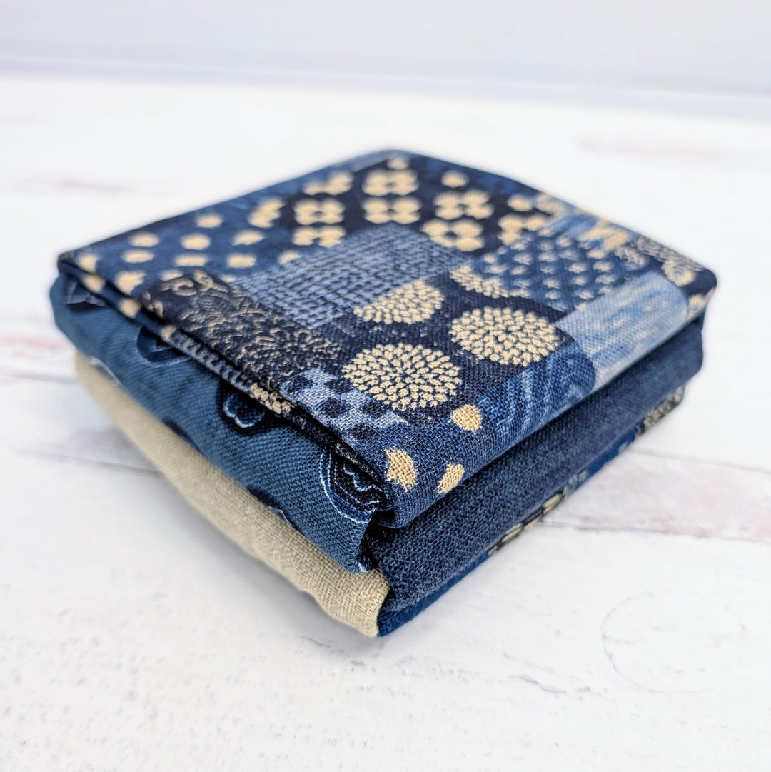 Stack of folded fabric pieces in blue, navy, and beige tones featuring subtle patterns, representing the Homespun Rustic Weave Fat Quarter Bundle for crafting and needlework projects.