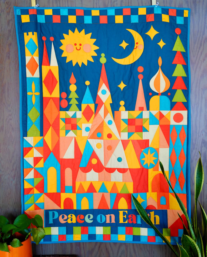 Peace on Earth Whole Cloth Quilt Kit featuring a colorful geometric cityscape design with sun, moon, stars, and trees, showcasing vibrant modern needlework in warm tones.