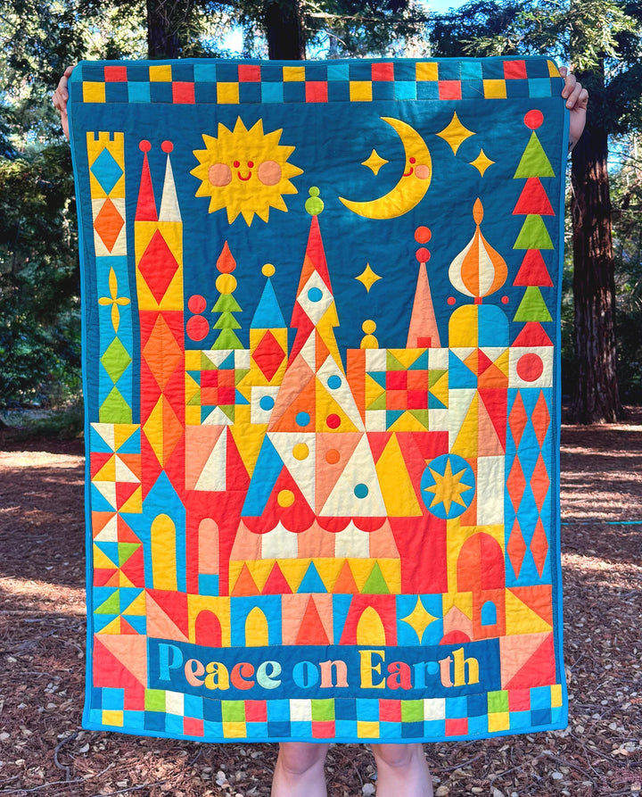 Person holding a brightly colored quilt featuring geometric shapes, a sun, moon, stars, trees, and the text Peace on Earth, showcasing the Peace on Earth Whole Cloth Quilt Kit design.