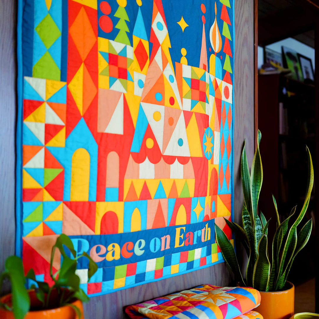 Brightly colored geometric patchwork quilt hanging on a wall with "Peace on Earth Whole Cloth Quilt Kit" text, surrounded by houseplants and folded fabric in a cozy craft room setting