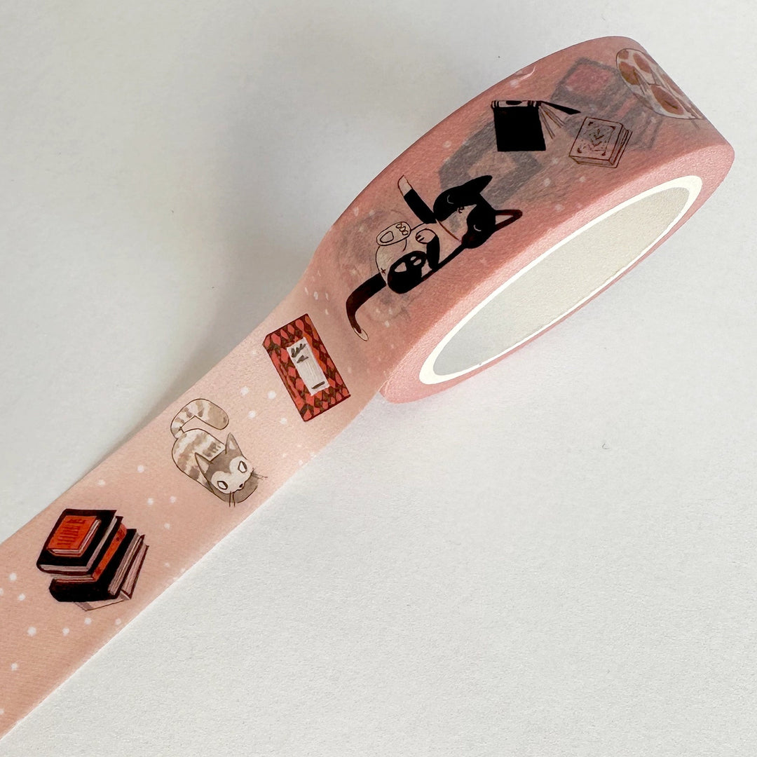 A roll of pink washi tape with cute illustrations of cats, books, and stamps, displayed diagonally on a light gray background.