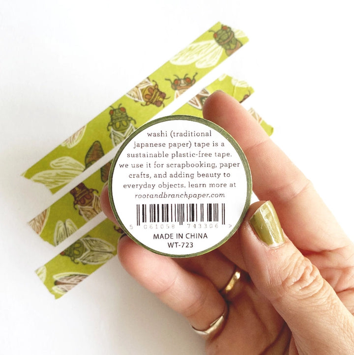Hand holding a roll of Cicadas Washi Tape with green cicada illustrations on a light olive background, next to strips of the tape unrolled on a white surface, showcasing Japanese craft supplies style.