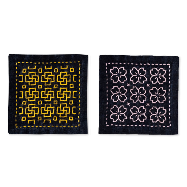 Two navy fabric coasters featuring geometric and floral patterns stitched with yellow and light pink embroidery thread, part of the Beginner Hitomezashi Sashiko Coaster Kit - Navy (SK-480).