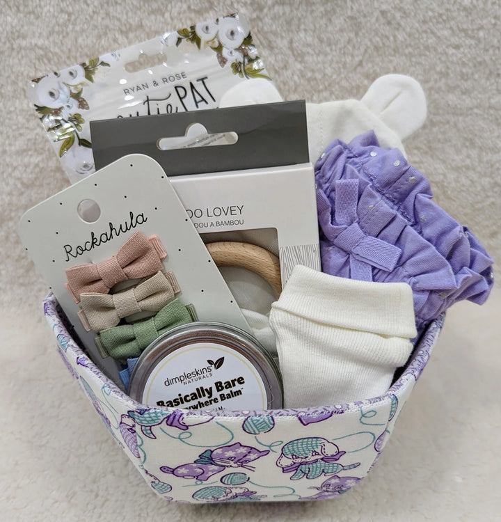 Fabric basket with a pastel pattern holding baby care items including socks, hair bows, balm, and a teething ring, showcasing the English Paper Piecing Bowls Pattern - The Goodness Bowl design.