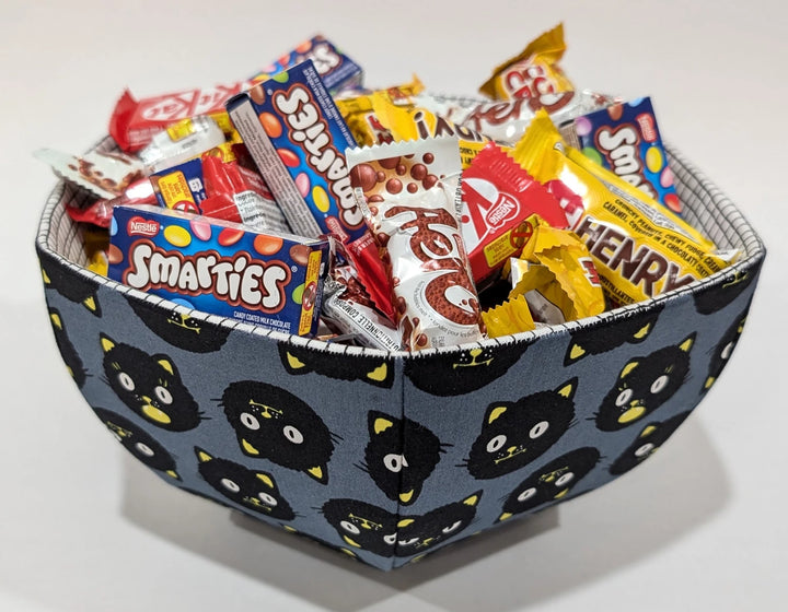 Fabric bowl with black cat faces pattern filled with assorted candies, showcasing the English Paper Piecing Bowls Pattern - The Goodness Bowl for modern needlework and craft enthusiasts.