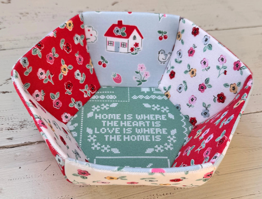 Fabric bowl made with colorful floral and house prints, featuring a cross stitch style center with the phrase "Home is where the heart is, love is where the home is," crafted using the English Paper Piecing Bowls Pattern - The Prosperity Bowl.