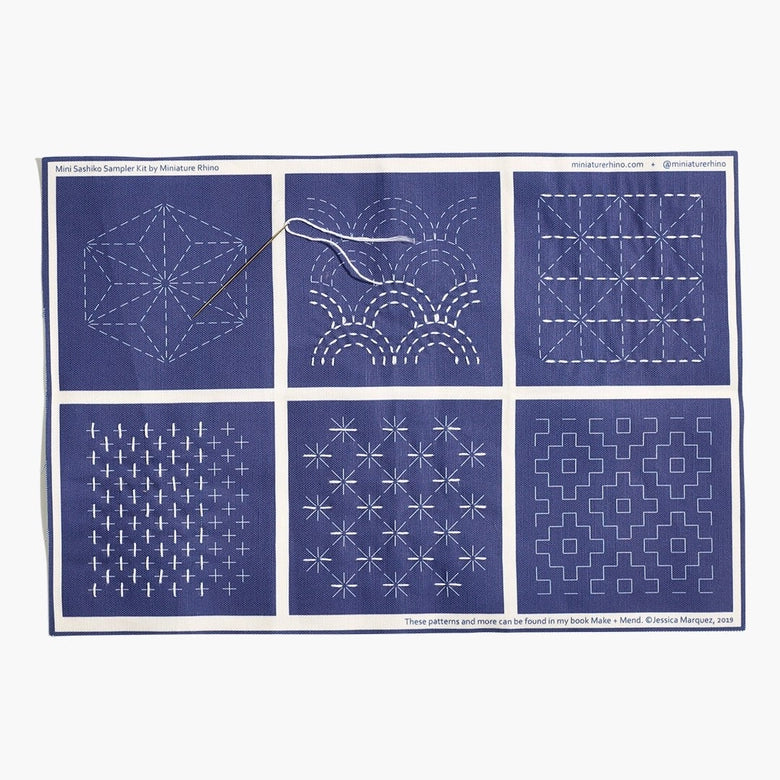 Sashiko Mini Sampler Kit (No. 1) featuring six traditional white sashiko stitching patterns on indigo fabric, including a threaded needle for practicing Japanese needlework techniques.