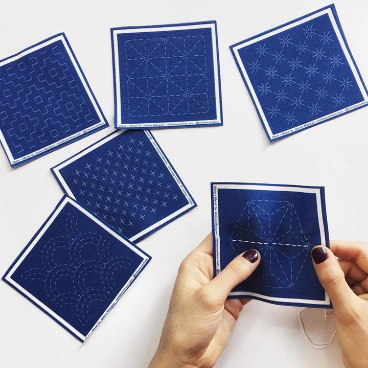 Hands holding a blue fabric square from the Sashiko Mini Sampler Kit (No. 1) with white stitched traditional Japanese embroidery patterns and five other stitched fabric squares laid out on a white surface.
