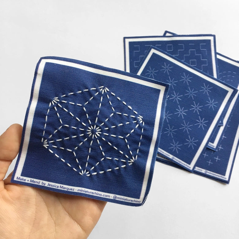 Hand holding a blue fabric square featuring white geometric sashiko stitches with additional blue fabric squares showing traditional sashiko patterns in the background, Sashiko Mini Sampler Kit (No. 1)