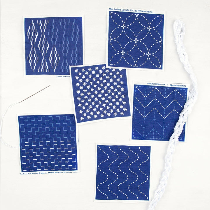 Sashiko Mini Sampler Kit (No. 2) displaying six blue fabric squares with white traditional Japanese Sashiko embroidery patterns, a threaded needle, and white embroidery floss.