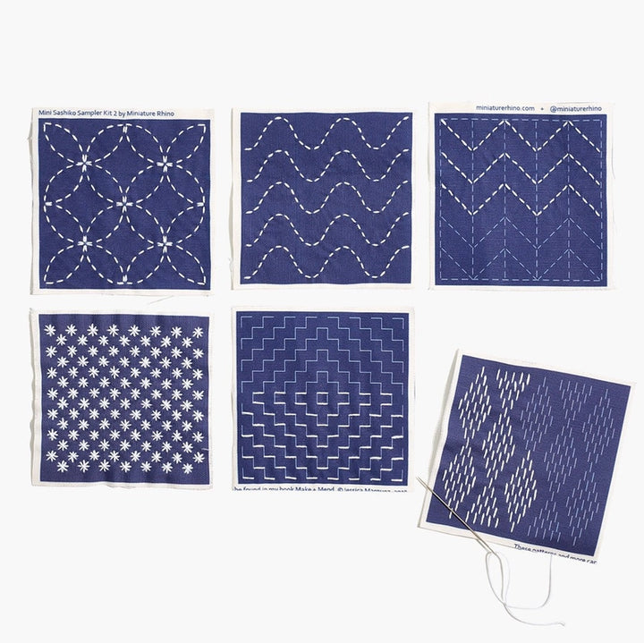Collection of six small navy blue fabric pieces showcasing different white sashiko embroidery patterns with needle and white thread, part of the Sashiko Mini Sampler Kit (No. 2)