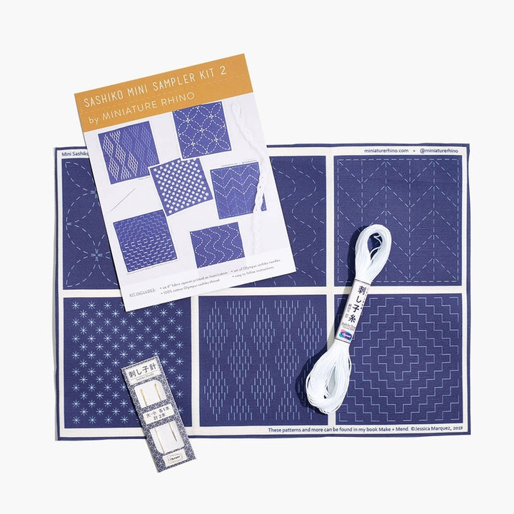 Sashiko Mini Sampler Kit (No. 2) featuring a navy blue fabric with white traditional sashiko embroidery patterns, a bundle of white embroidery thread, and needles for hand stitching.