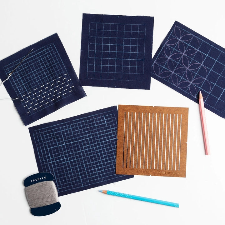One-Quarter Inch Sashiko Grid Stencil (Oil-Board) shown with navy fabric pieces featuring various grid and sashiko stitch patterns, a pencil, and gray embroidery thread for modern needlework.
