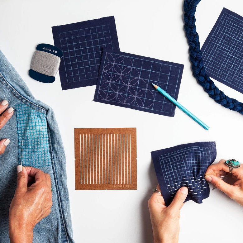 Hands holding indigo fabric marked with a grid and circular designs alongside a brown One-Quarter Inch Sashiko Grid Stencil (Oil-Board), thread, and a denim jacket being prepared for sashiko stitching.