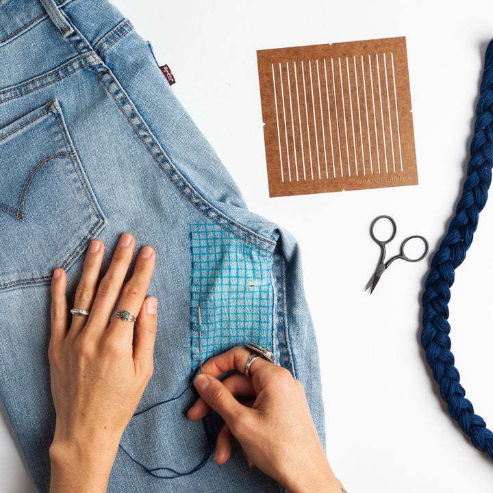 Hands using the One-Quarter Inch Sashiko Grid Stencil (Oil-Board) to create precise sashiko stitching on light blue denim fabric, with embroidery scissors and blue embroidery thread nearby.