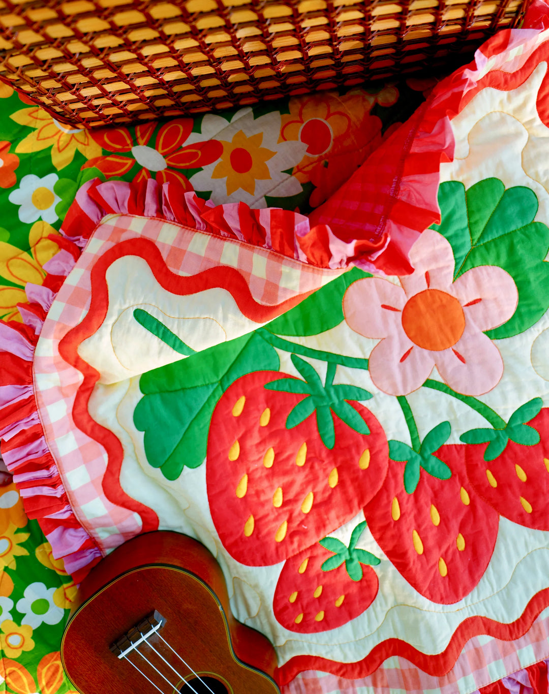 Bright strawberry and flower patterned quilt with red ruffled edges, laid on a colorful floral background with a small wooden ukulele, featuring the Strawberry Whole Cloth Quilt Kit design.