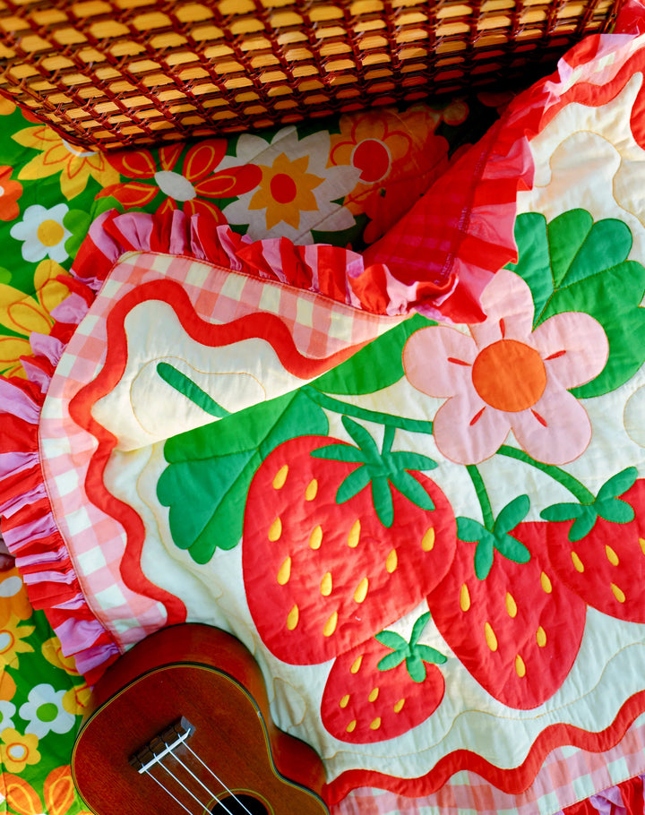 Bright strawberry and flower patterned quilt with red ruffled edges, laid on a colorful floral background with a small wooden ukulele, featuring the Strawberry Whole Cloth Quilt Kit design.