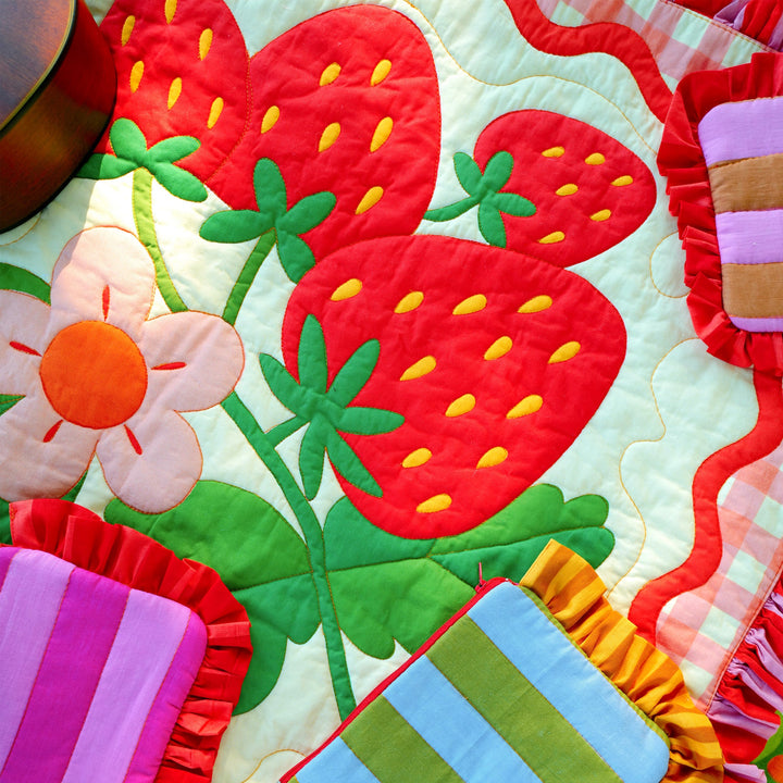 Close-up of a colorful quilt with large red strawberries and green leaves, surrounded by pillows, showcasing the Strawberry Whole Cloth Quilt Kit with vibrant hand-stitched details.