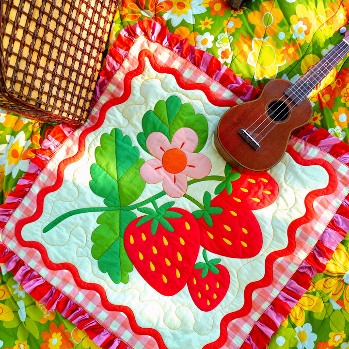 Colorful quilted pillow featuring large red strawberries and green leaves with a pink flower, bordered by red ruffle, displayed with a ukulele and wicker basket, showcasing the Strawberry Whole Cloth Quilt Kit.
