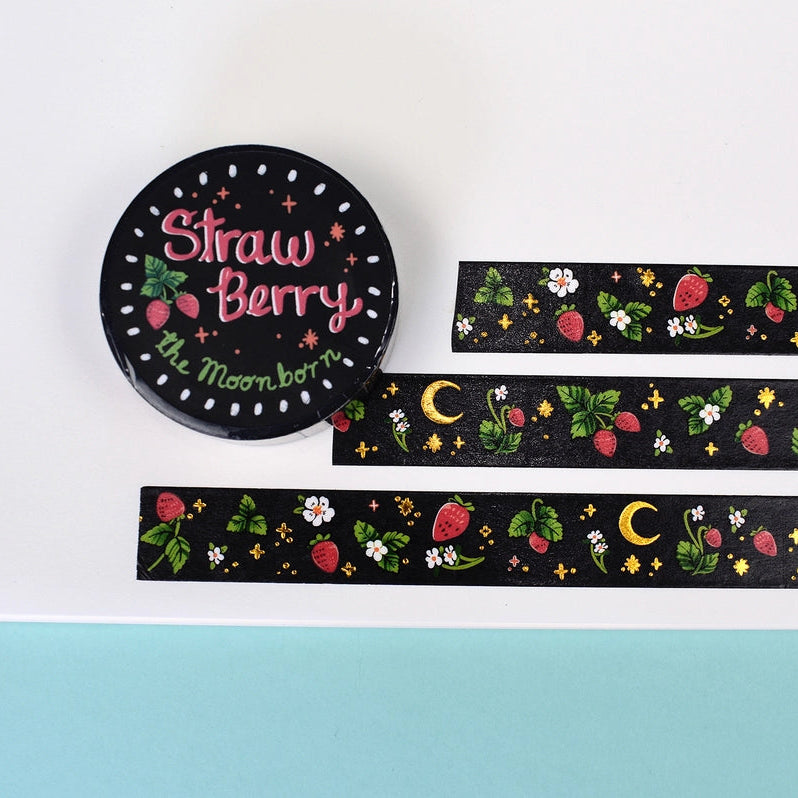 A round black sticker or coaster with the words 'Straw Berry' and 'by Moonborn' in pink and green, shown beside two narrow black patterned strips featuring strawberries, green leaves, white flowers, and yellow crescent moons, arranged on a white board against a light blue background.