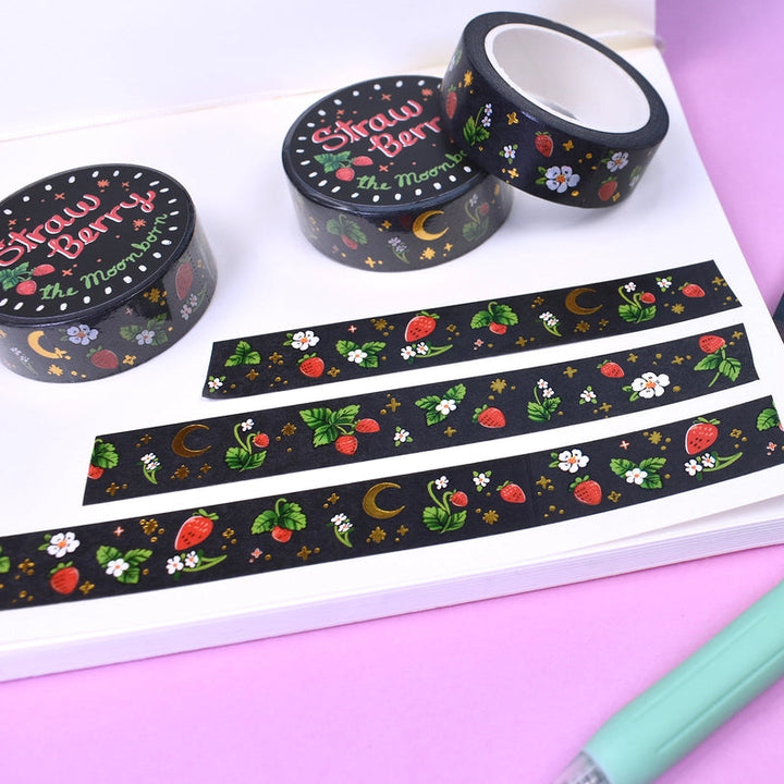 A flat-lay of Strawberry Moon washi tape products on a pink background, including rolls, strips, a notepad, and a mint green pen.