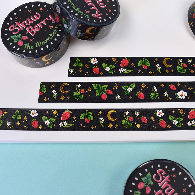 Black washi tape with a strawberry and moon pattern, shown both rolled up and unrolled on a white sheet over a light blue background.