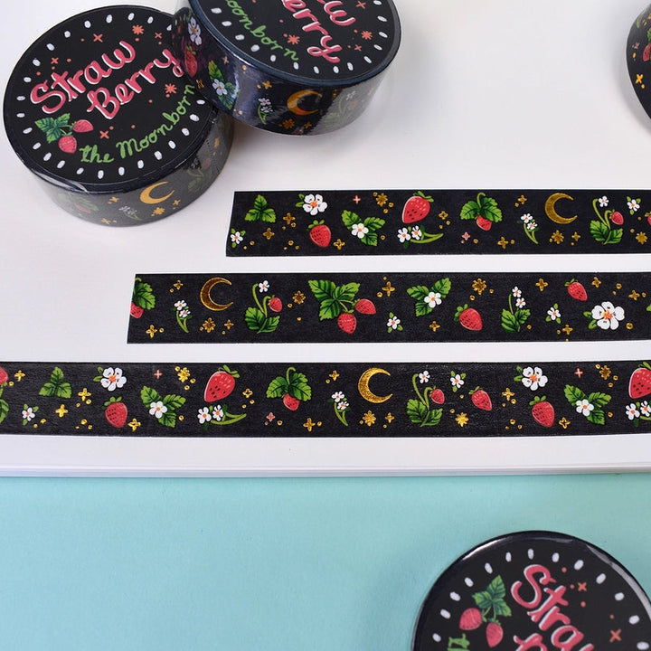 Black washi tape with a strawberry and moon pattern, shown both rolled up and unrolled on a white sheet over a light blue background.