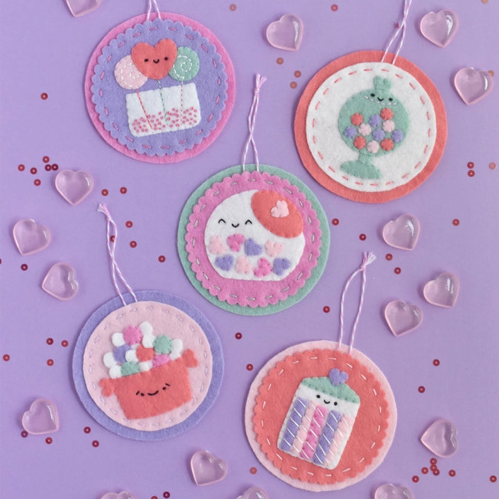Sweetheart Shoppe Ornament Kit featuring five colorful embroidered felt ornaments with smiling designs, displayed on a purple background with scattered heart-shaped accents.