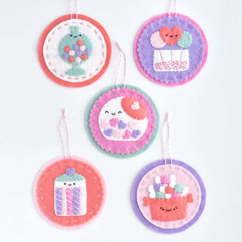 Sweetheart Shoppe Ornament Kit featuring five colorful felt ornaments with charming embroidered designs of a candy jar, heart balloons, a smiling dumpling, a striped cupcake, and a basket of yarn, each with hanging loops.