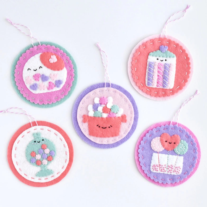 Five colorful felt holiday ornaments with smiling designs including a cake, a gift box with balloons, a candy jar, and a sewing spool, each crafted with embroidery floss and labeled Sweetheart Shoppe Ornament Kit.