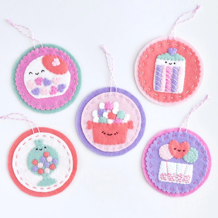 Five colorful felt holiday ornaments with smiling designs including a cake, a gift box with balloons, a candy jar, and a sewing spool, each crafted with embroidery floss and labeled Sweetheart Shoppe Ornament Kit.