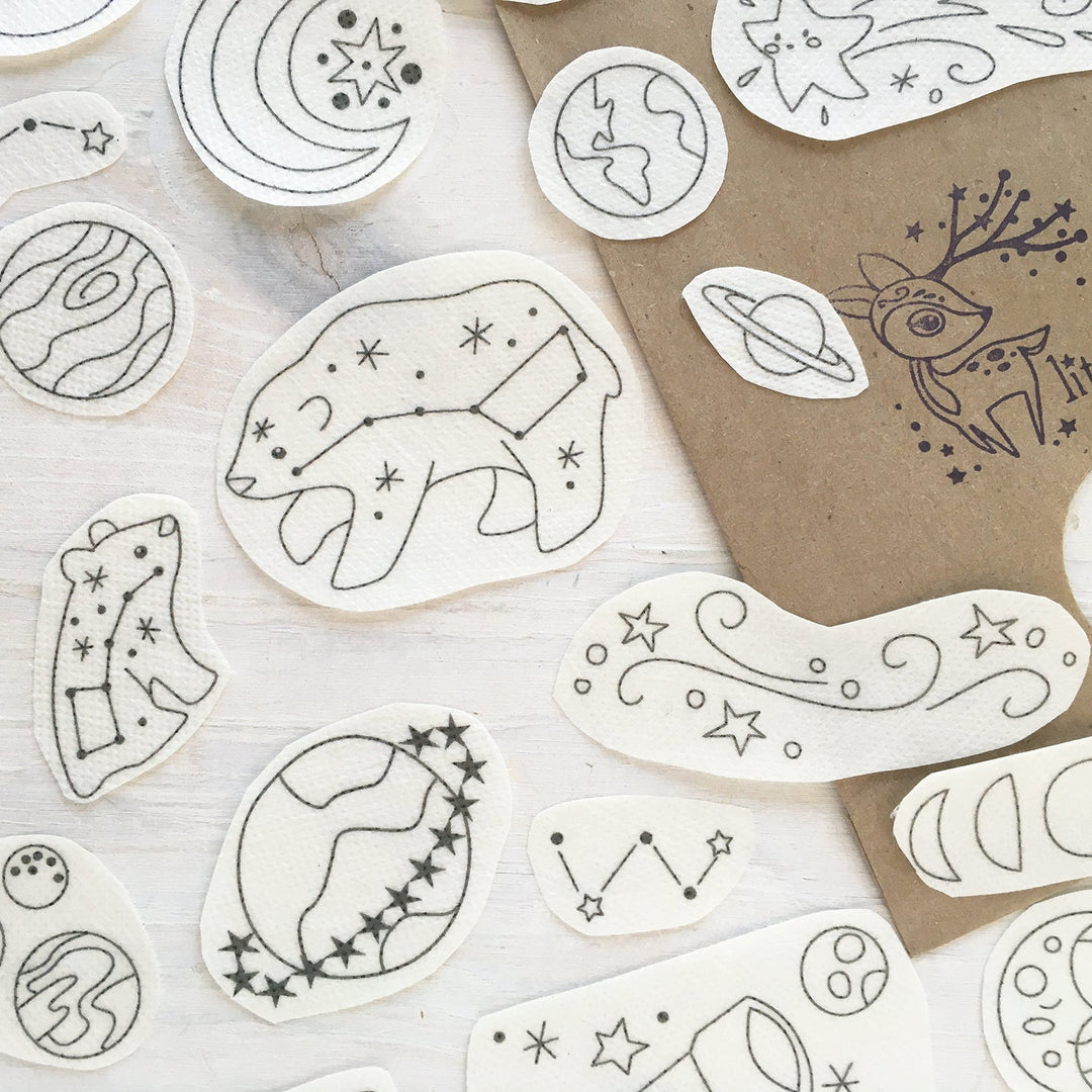 A collection of white paper cutouts featuring black line-art celestial and animal illustrations, arranged on a light wood surface next to a brown kraft paper bag printed with 'little deer'.