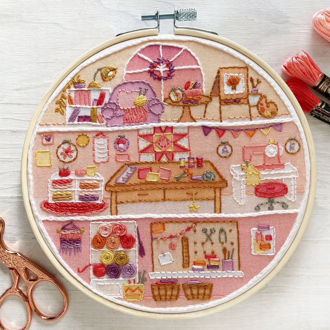 A circular embroidery hoop displays a detailed, multi-colored stitched design of a cozy sewing and crafting studio. The hoop is styled on a white wood background alongside a pair of rose gold embroidery scissors on the left and three skeins of pink and red embroidery floss on the right.