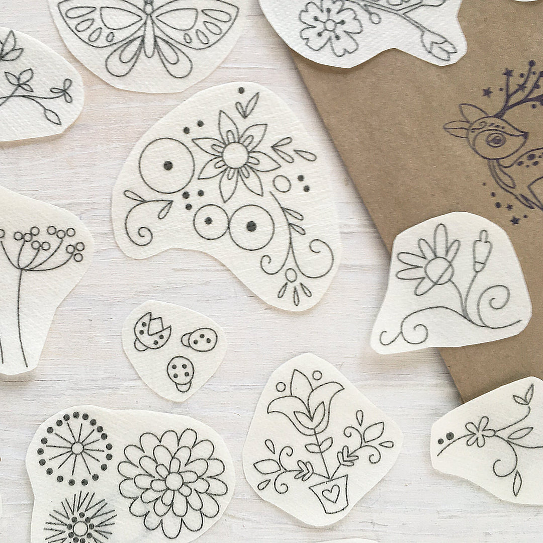 A flat-lay arrangement of white fabric patches with black hand-drawn botanical and insect line art, alongside a brown kraft paper sheet with a deer illustration, on a light wood background.