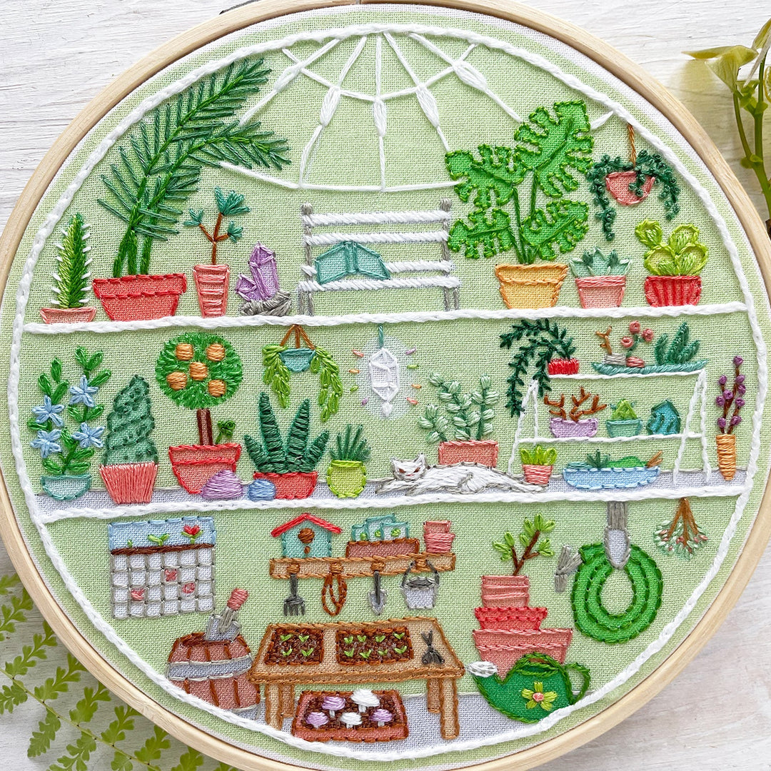 A circular embroidery hoop containing a colorful cross-stitch design of a greenhouse filled with potted plants, shelves, gardening tools, and small decorative elements, displayed on a white wood surface with greenery props.